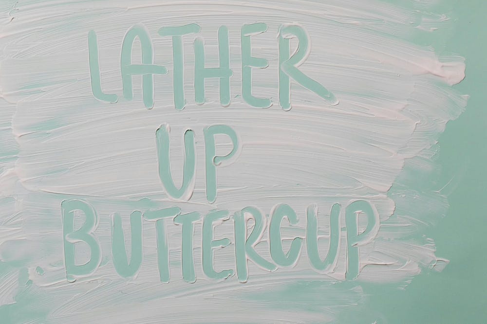 Teal background with white sunscreen smeard on it. In the sunscreen “lather up buttercup” has been written with a finger.