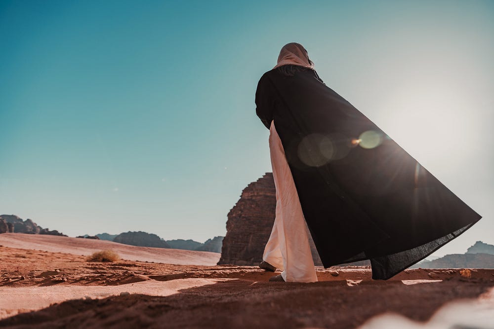 A woman standing alone in the desert on a sunny day with her head covered wearing a full length cloak.