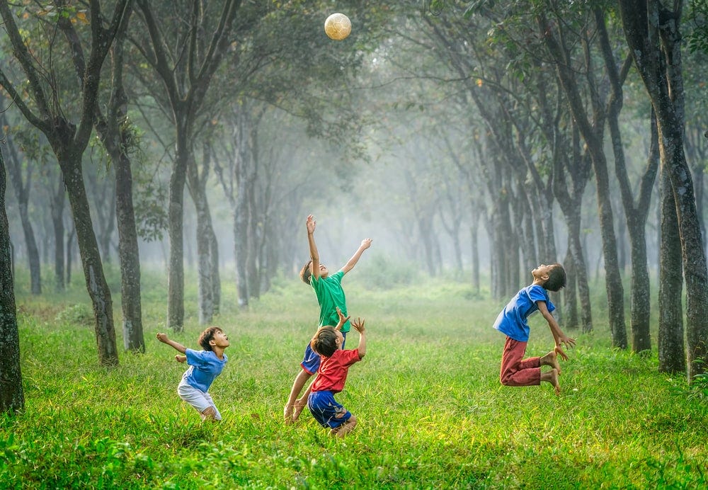 A dreamy image of four young children in colorful clothes jumping to catch a ball. They’re on a patch of spring-green grass between two columns of just-budding trees. There’s light fog in the background.