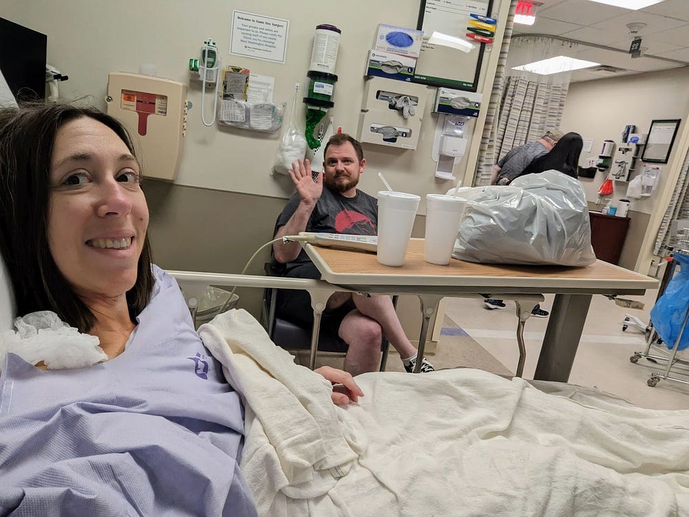 Author, Heather Gioia, in a hospital before going into surgery. Her partner is in the background waving. Heather has a nervous smile and is wearing a purple hospital gown while laying in a hospital bed with a blanket on.