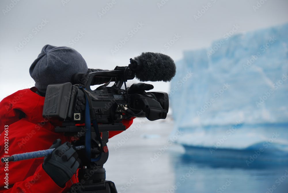 Cameraman wearing red coat, beanie, and gloves points a camera at an iceberg in Antarctica