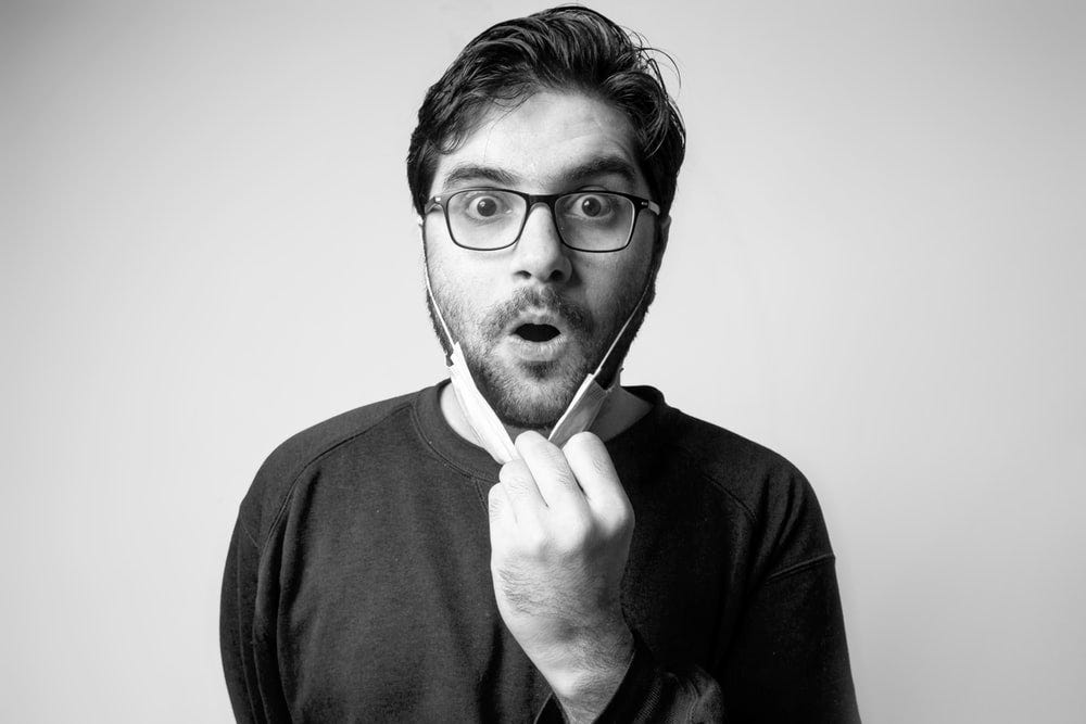 Grayscale picture with a dark-haired person with a beard, wearing glasses, who is pulling their mask down, looking surprised with an open mouth.