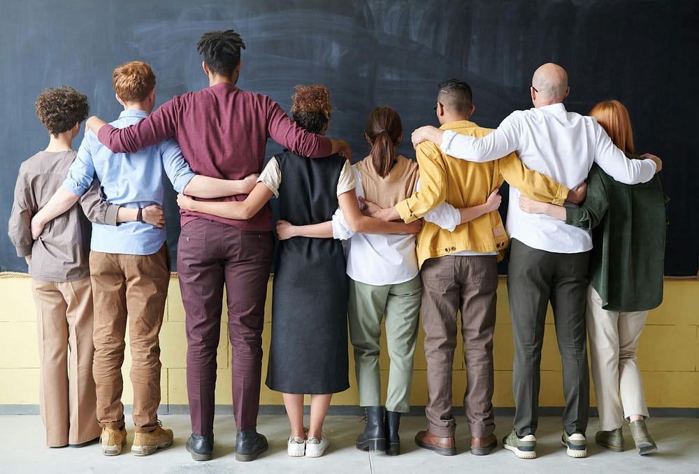Image of eight people standing with their arms around each other and their backs to the camera. They are standing in front of a wall that is a chalkboard on the upper half and light yellow on the lower part. The people vary in age, size, gender, and ethnicity.