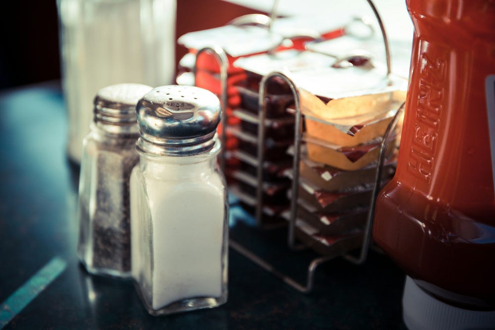 Close up of two clear glass salt and pepper shakers with chrome tops sitting on a table next to a metal sugar packet holder and red plastic ketchup bottle.
