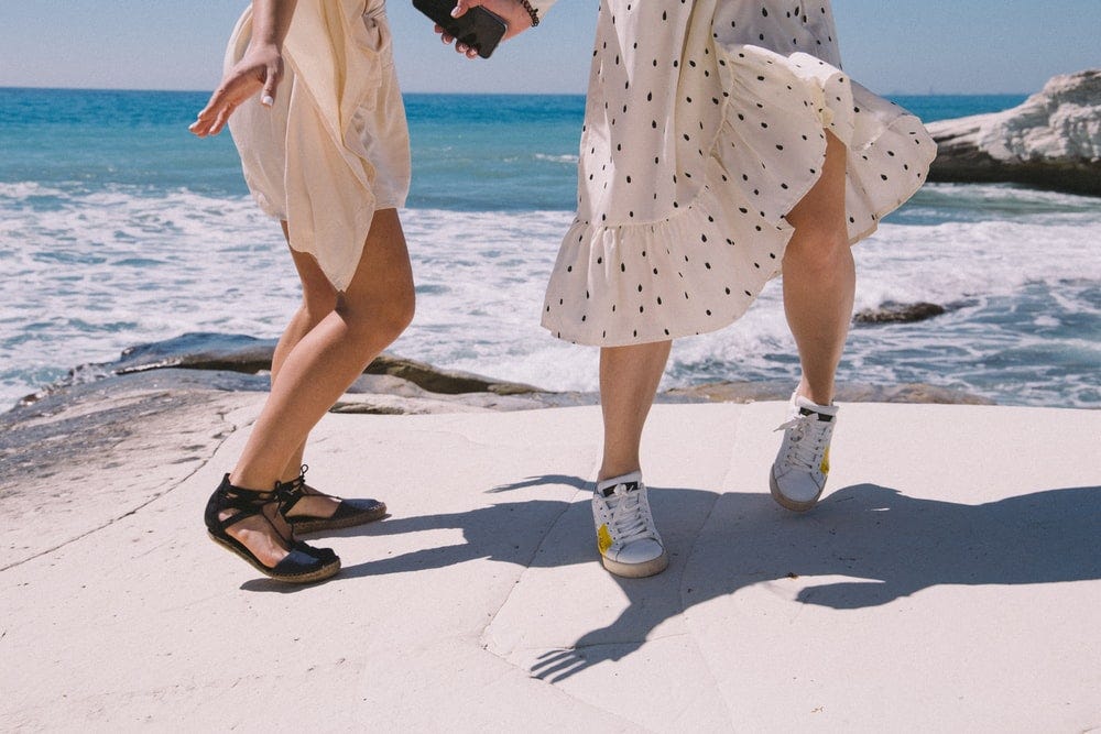 women in yellow and white polka dots dress and white shoes standing side by side jumping happily; the sea behind them