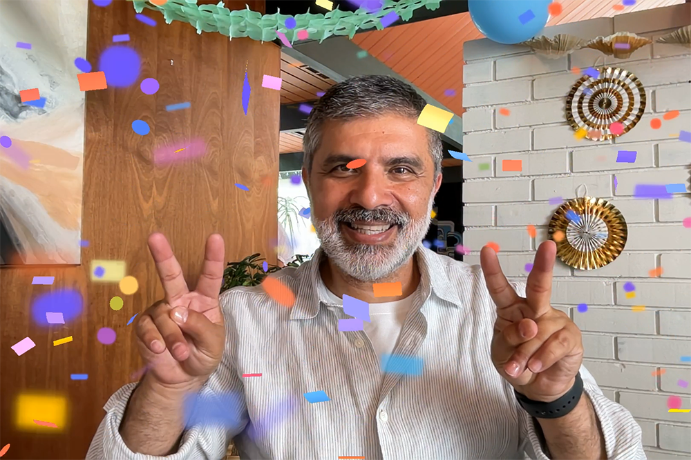 Digital confetti floats in front of a man holding up both hands with the peace sign.