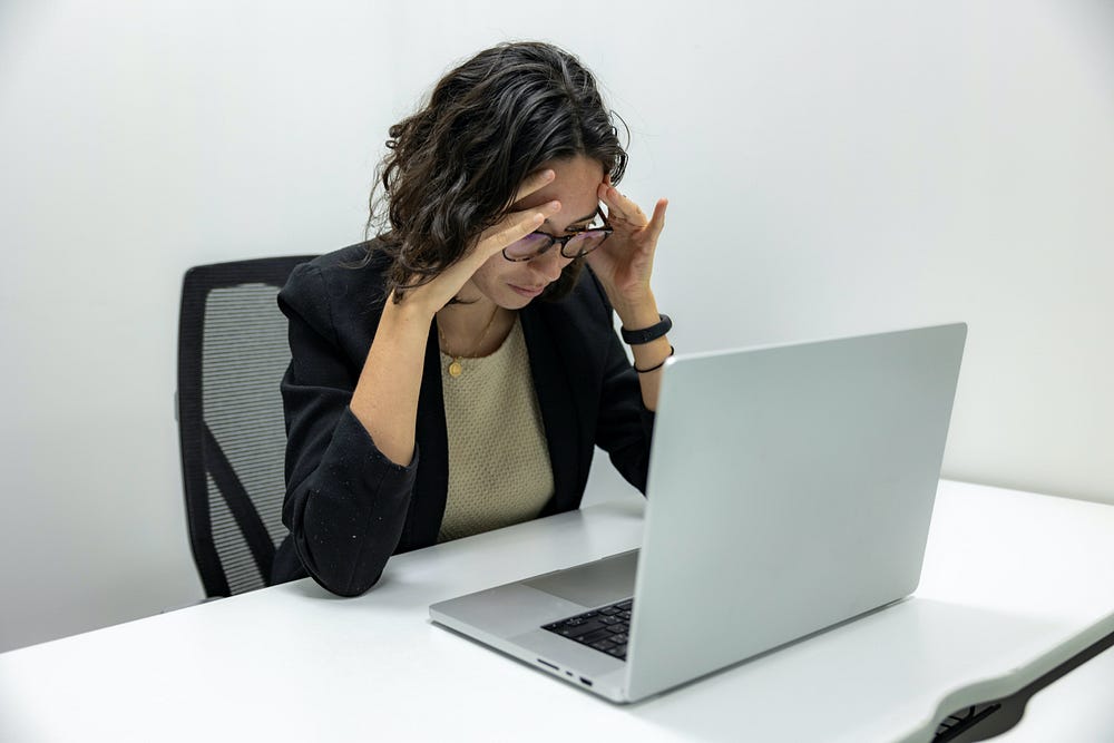 A young woman wearing glasses sits at a table looking at a laptop. Her elbows are bent resting on the table, and her fingers are placed on her temples.