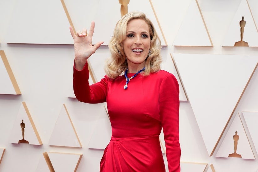 Marlee Matlin is walking the Oscar’s red carpet. She is standing, wearing a red dress and signing “I love you” and smiling.