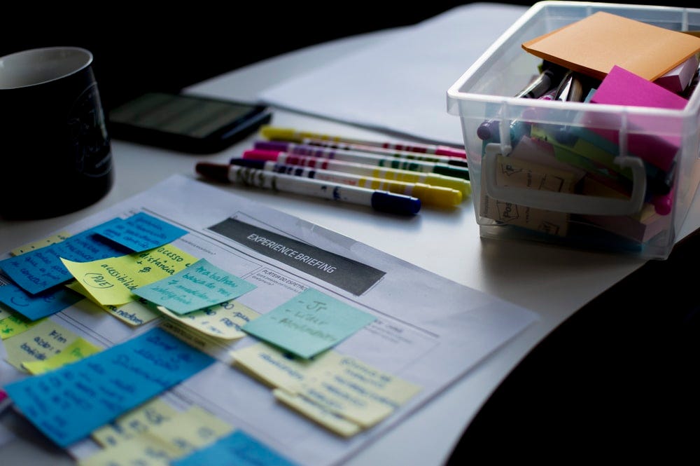 Multicoloured felt-tip pens sitting on a white table with an A4 piece of paper covered in post it notes and a box of random stationary