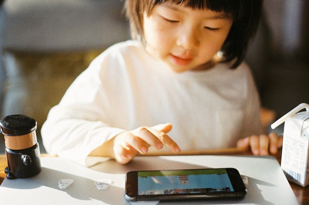 Criança pequena de cabelos escuros na altura dos ombros e blusa branca. Ela interage com um smartphone que está sobre uma mesa. Na tela do dispositivo vemos uma ilustração infantil.