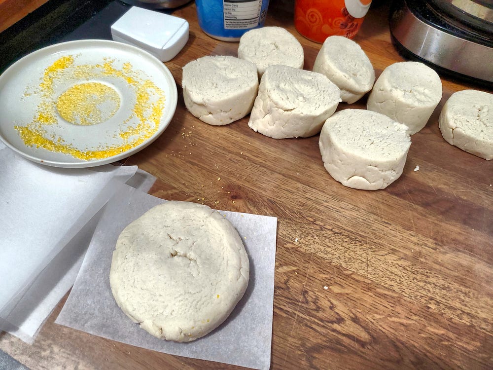 Seven pieces of dough sitting on a clear plastic cutting mat with a small saucer with cornmeal in it to the left of the dough. In the bottom of the photo is a flattened piece of dough in a circular shape sitting on a piece of parchement paper.