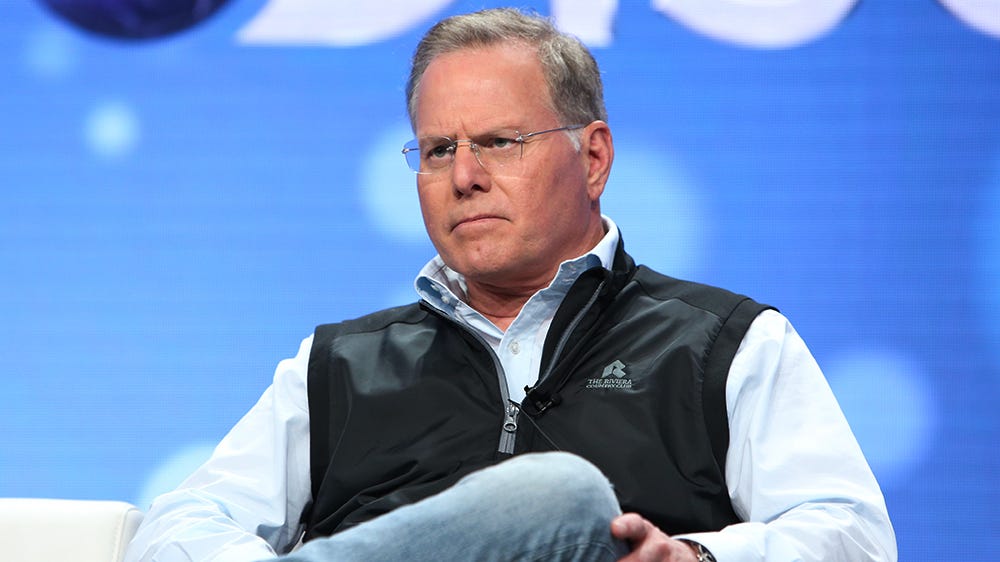 A picture of David Zaslav, Discovery’s CEO, sitting in a chair with his legs crossed. He is wearing glasses and looking forward with a neutral smile. He is wearing a white dress shirt with a black half-zip vest and wearing blue jeans.