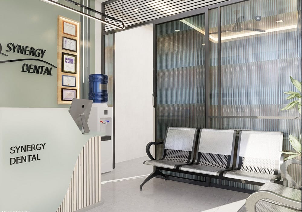 A modern dental clinic reception interior at Synergy Dental featuring a sleek curved desk with a mounted computer, framed certificates on a soft green wall, and a water dispenser. The waiting area includes black metal chairs with mesh seating beside large glass partitions that bring in natural light. The clean, minimalistic design combines comfort, functionality, and aesthetics to create a welcoming dental clinic atmosphere.