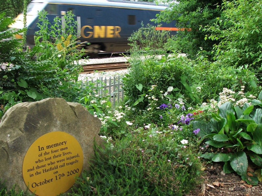 A small memorial garden in front of a railway where a train passes. The memorial reads: “In memory of the four men who lost their lies, and those who were injured, in the Hatfield rail tragedy on October 17th 2000”