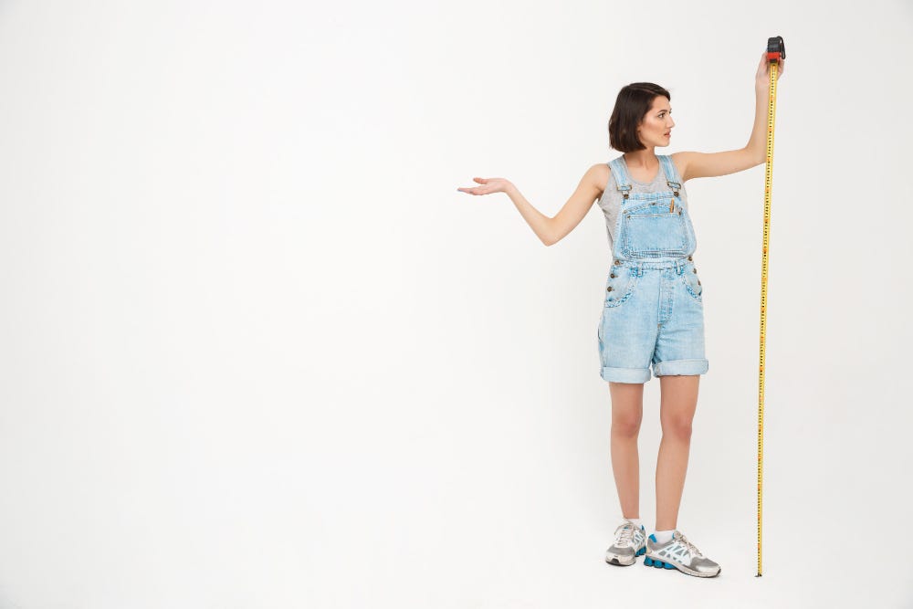 A woman with pale skin holds a tape measure to measure her own height, with a shrugging gesture.