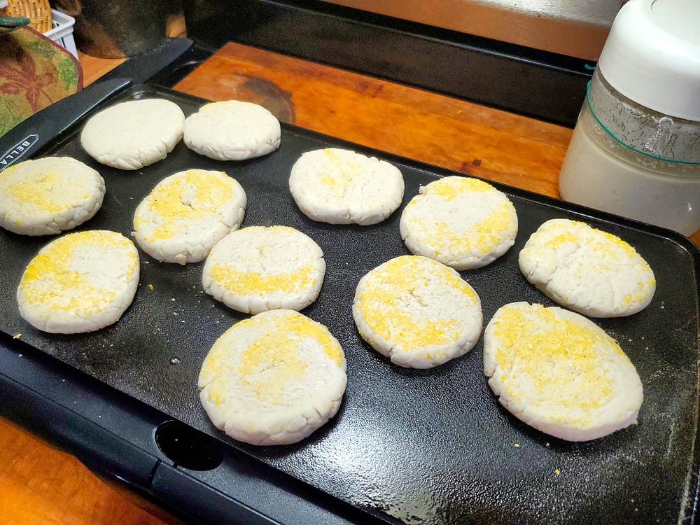 Twelve English muffins, some with cornmeal on the top, sit on a black table top griddle. Just behind the griddle on the right is a glass container of sourdough starter.