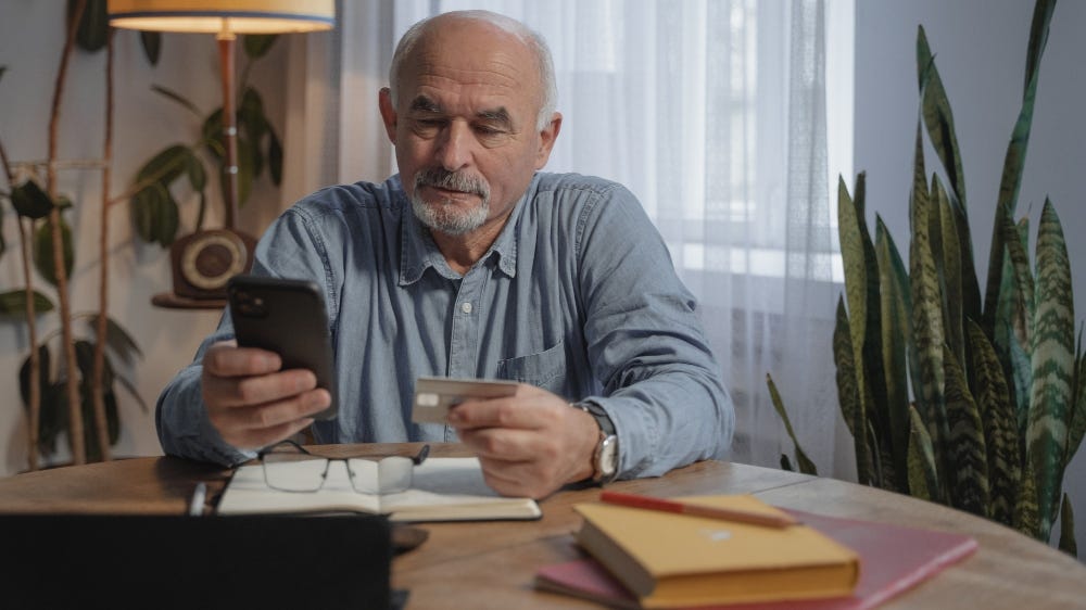 Foto de um homem idoso segurando um celular em uma mão e um cartão de crédito em outra.