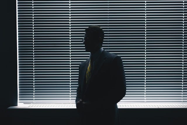 The silhouette of a suited man stands in front of a window with blinds, casting a shadow in the softly lit room.