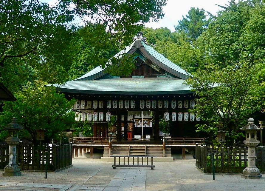 Japanese shrine with green curved roof and many hanging white lanterns beneath its eaves. Two stone lanterns flank the sides. Green leafy trees stand behind and beside the shrine.