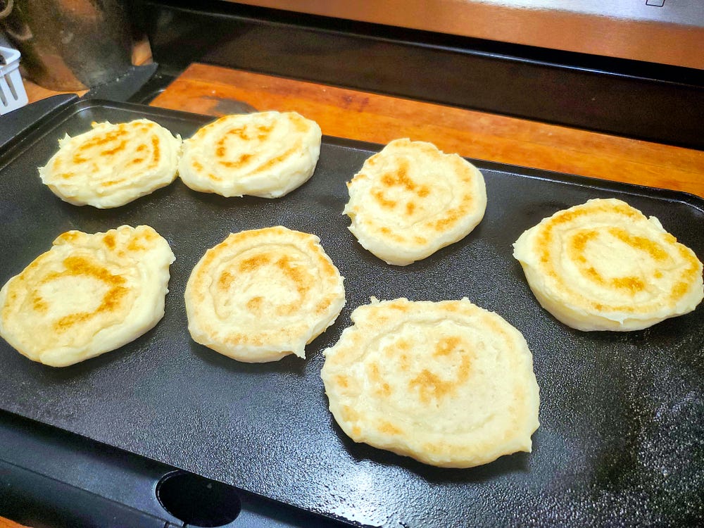 Seven free form English muffins with golden griddle marks sitting on a black table top electric griddle.