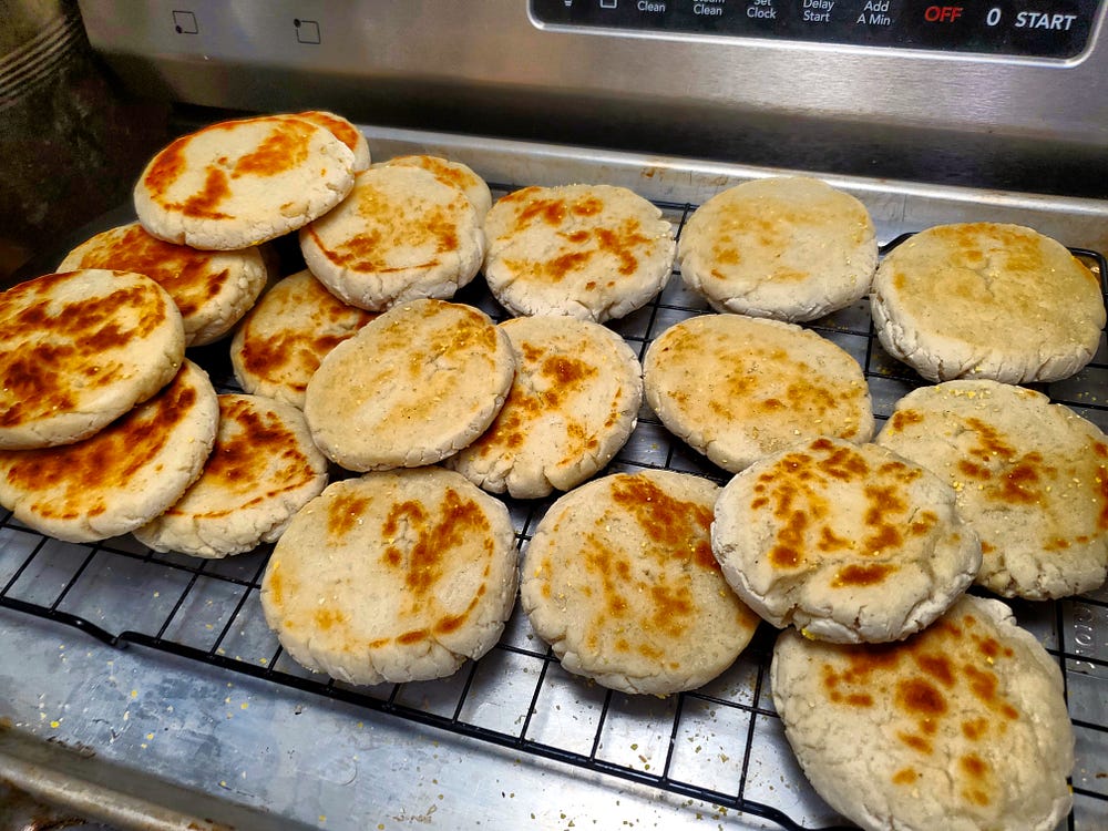 Twenty four English muffins sit cooling on a wire rack set over a rimmed cookie sheet.