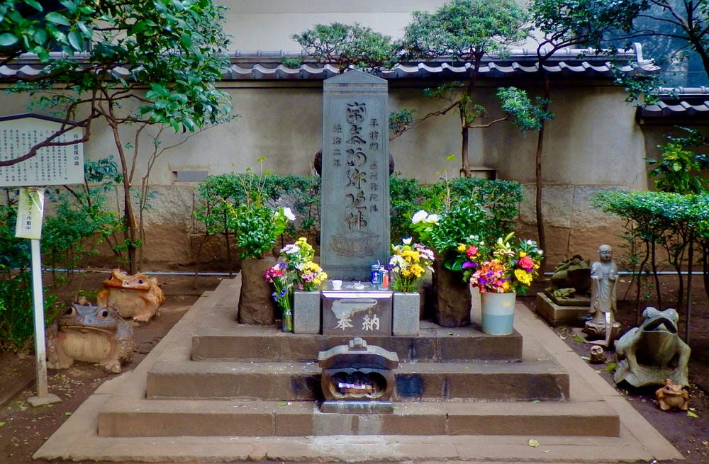 Stone monument with buckets and pots holding flowers in front. Frog statues and one Buddhist statue on either side. Trees and bushes in back.