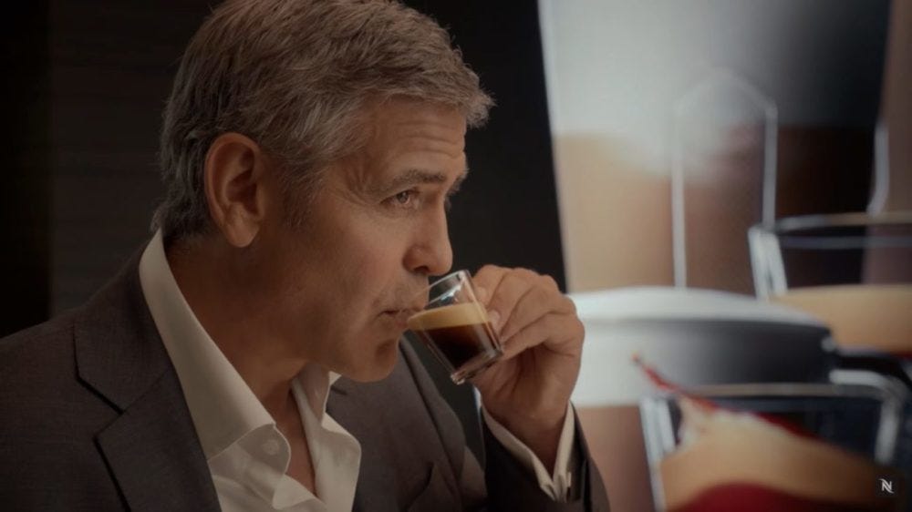 George Clooney vestido de terno tomando uma xícara de café, olhando para o horizonte, reflexivo