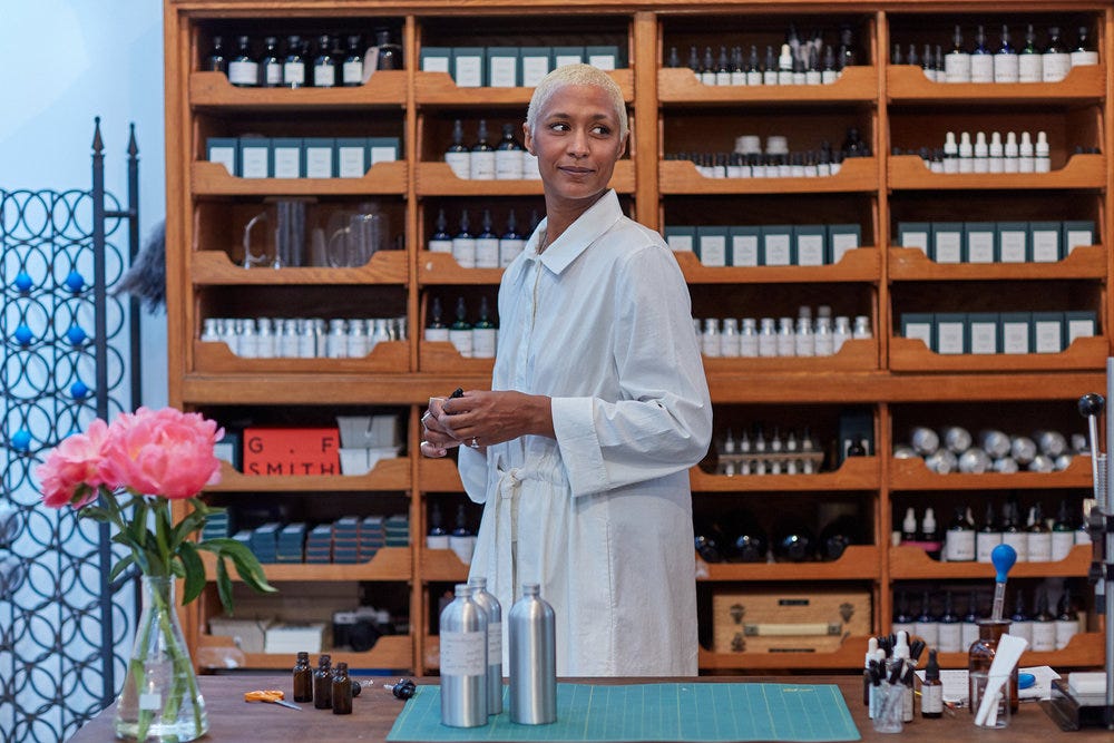 Maya stands in front of bookcase, filled with glass bottles, in a white lab coat. Some pink flowers sit in a vase on her left