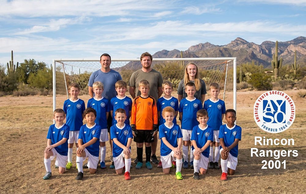 A group portrait of the Rincon Rangers 2001 youth soccer team in blue and white uniforms, standing in front of a soccer goal with a desert landscape and rugged mountains in the background.