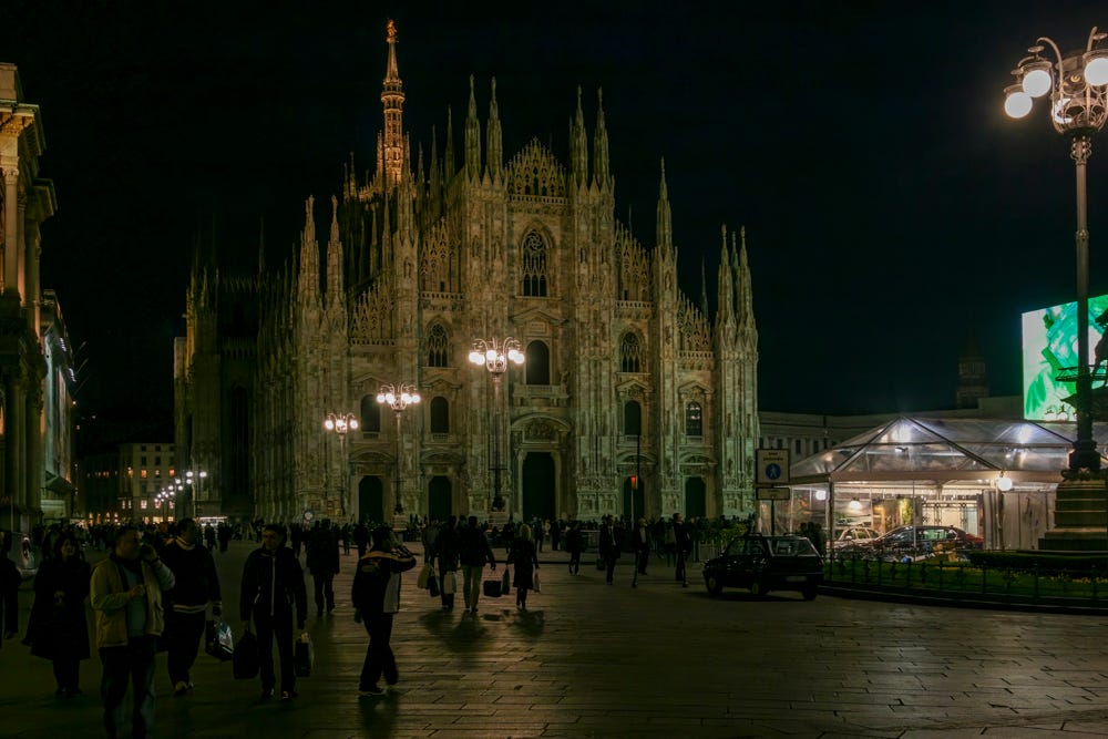 Nighttime view of Milan Cathedral with silhouettes of 50–60 people walking in Piazza del Duomo. Four lampposts with globe lights provide most of the lighting.