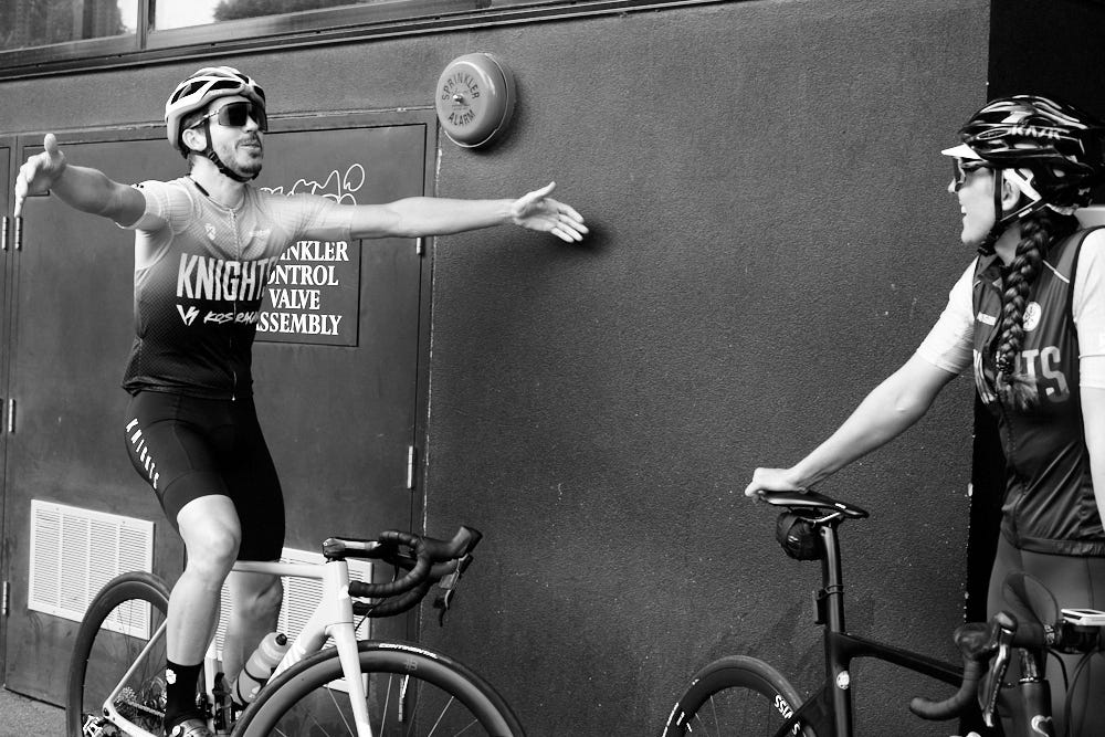 A man and woman on bicycles. Man holding out his arms for a hug. Woman laughing.