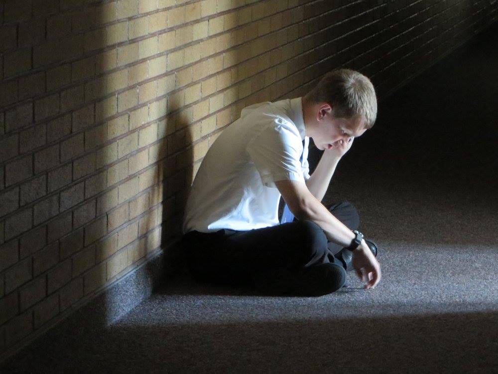 Young man talking on the phone, sitting against a wall, a ray of light from a doorway falling upon him.
