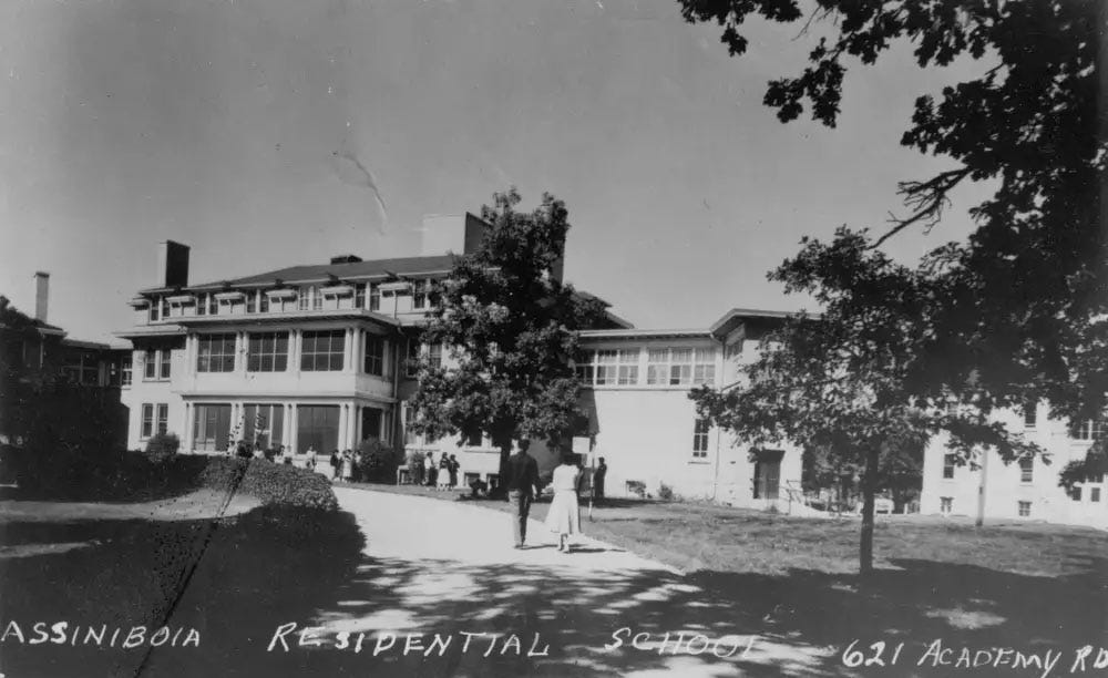 Black and white picture of thethree-story Assiniboia Residential School surrounded by trees, a road, a hedge, and a manicured lawn. Two people are walking towards the school, holding hands.