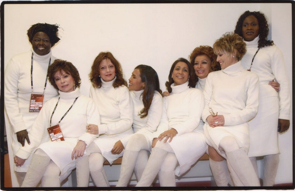 Eight women of different races and nationalities, are wearing all white and sitting on a white sofa.