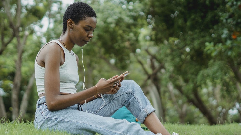 Fotografia de uma mulher sentada em um gramado. Ela está com um fone em um dos ouvidos e mexe em seu celular.