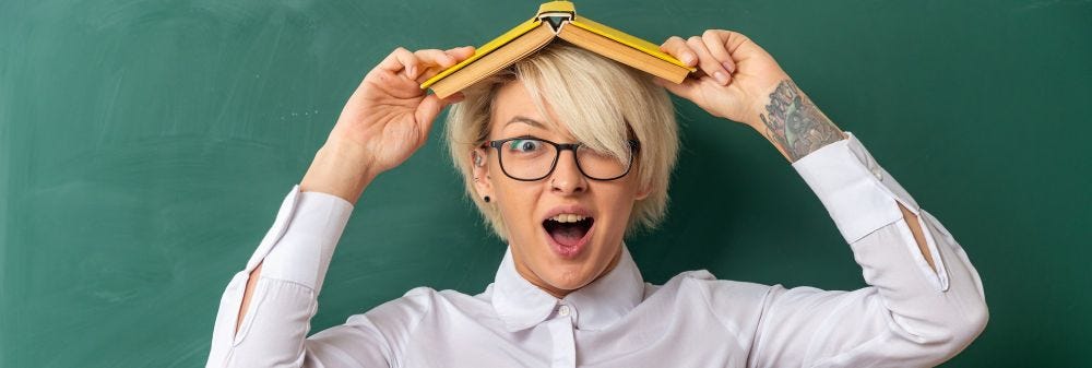 Excited young blonde female teacher wearing glasses in classroom standing in front of chalkboard holding book on head looking at camera
