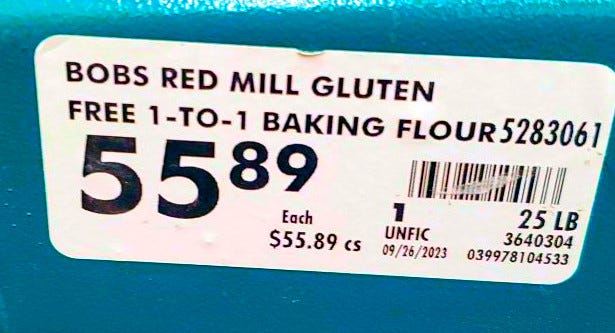 A white tag on a blue green background with the words Bobs Red Mill Gluten Free 1 to 1 Baking flour and a price of $55.89 each. There are other numbers and a barcode on the lable.