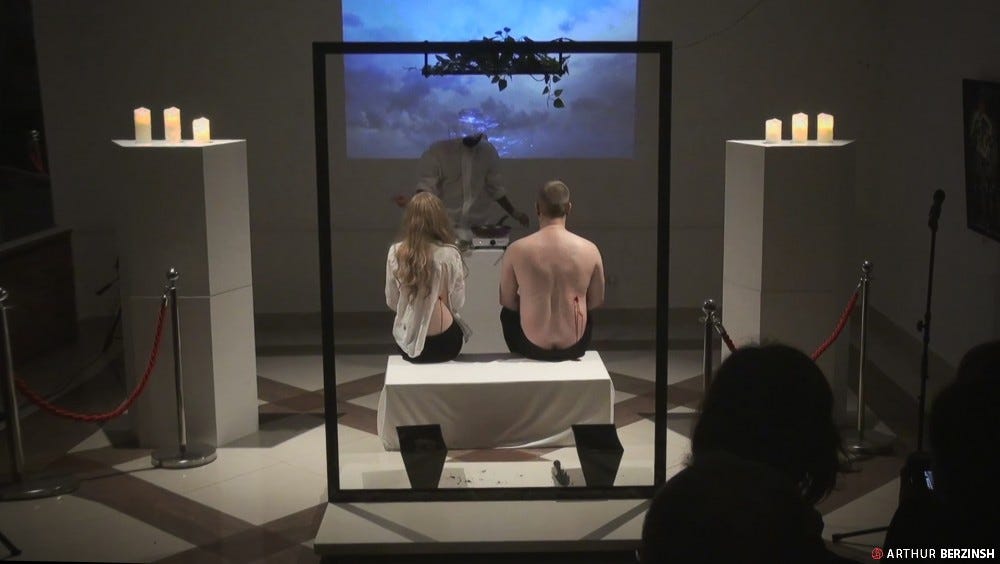 A photo of Eschatology by Arthur Berzinsh. Two performers, a woman and a man, sit with their backs to the audience. The man is shirtless. The woman has a slit in the back of her shirt. Both performers have a bleeding cut on the right side of their backs. The performers face a woman wearing srubs who is frying their flesh in a pan. They are surrounded by candles on pedestals and there is a photo of a thunderstorm on the wall behind them.