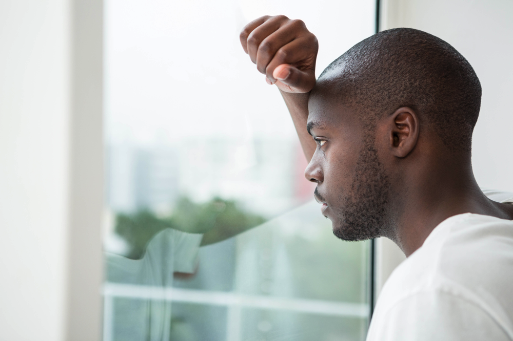 A young black man peers out the window thoughtfully with his first raised above his head.