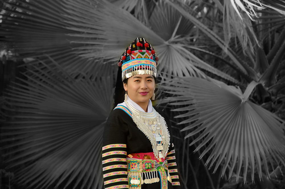 The author Jer Xiong wears her traditional Hmong clothes. She is in color while her background of a large plant is in black and white.