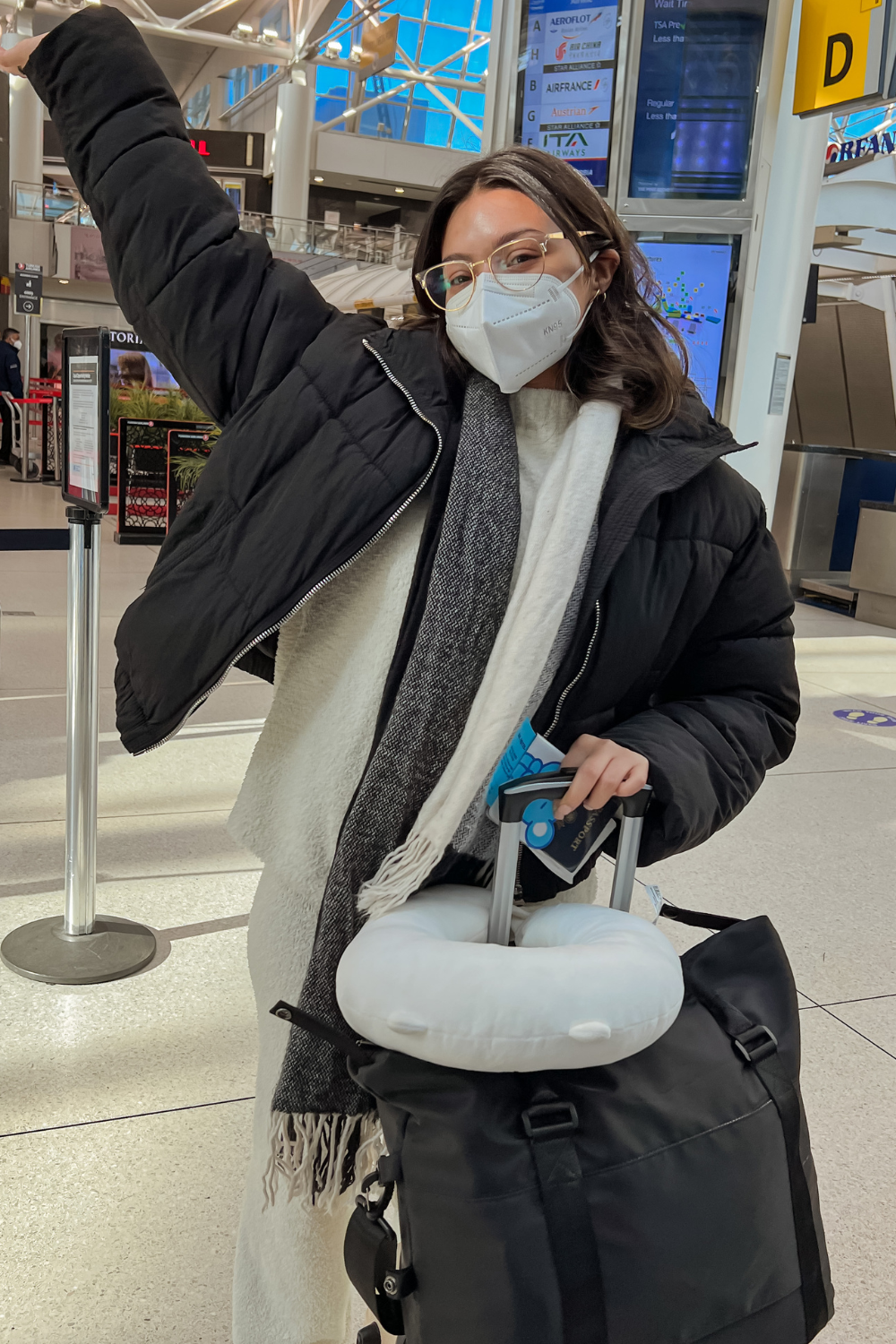 Woman with winter clothes and a mask on is in an airport holding her luggage and passport.