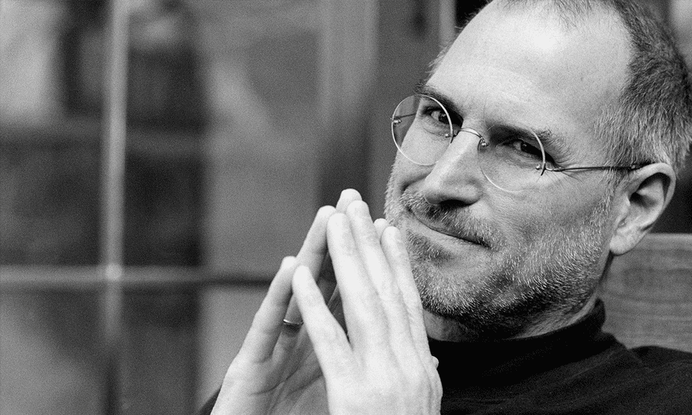 Foto em preto e branco fundo desfocado a frente Steve Jobs, homem branco, de óculos sorrindo com as pontas dos dedos unidas frente ao rosto.