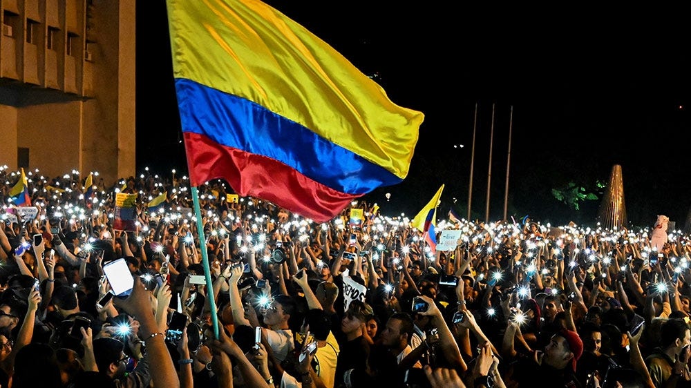 People protesting at the streets, holding their cellphones up. One big Colombian flag in the front.