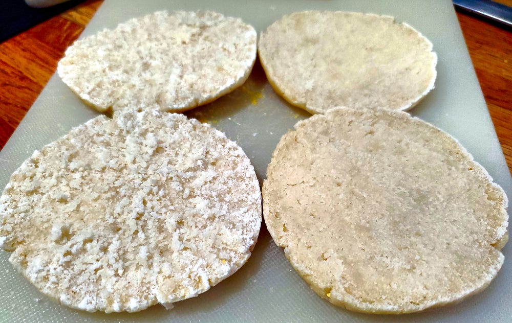 Two English muffins sit on a cutting board. They have been split open. The Two halves on the right have been sliced with a knife, showing a smooth interior. The halves of the left have been split open with a fork and show a rough texture with nooks and crannies.