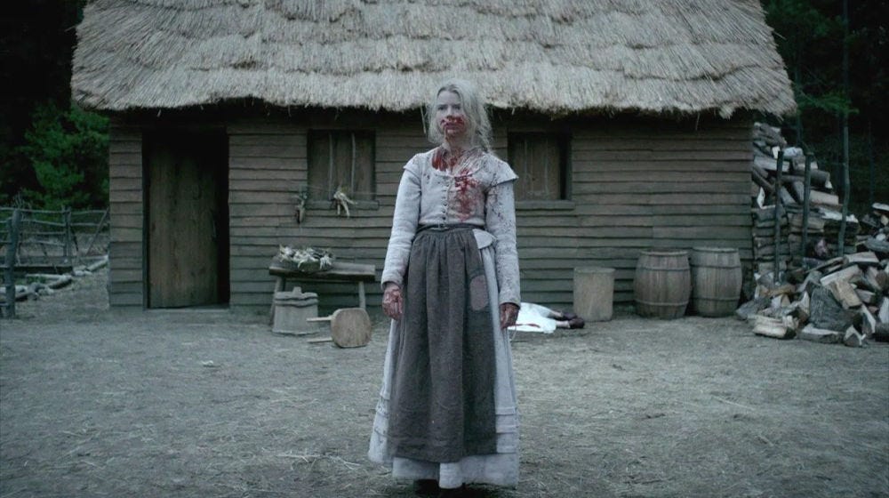 A woman dressed in 17th century clothing covered in blood stands in front of a wooden cabin