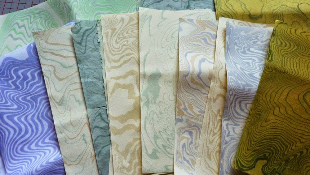 Patterned marble papers in shades of purple, green, and beige are arranged in a fan. Waves and swirls create a dynamic, artistic texture.
