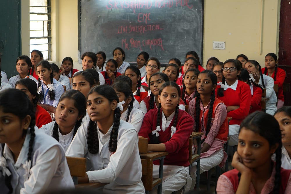 a classroom full of female students in a white uniforms with some of them wearing a red sweater. most of them are wearing plaits in their hair with white ribbons to tie them.