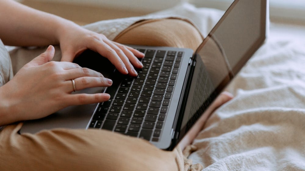 Foto de uma pessoa digitando em seu notebook. O enquadramento mostra apenas suas pernas cruzadas, o notebook sobre elas e suas maõs sobre o teclado.