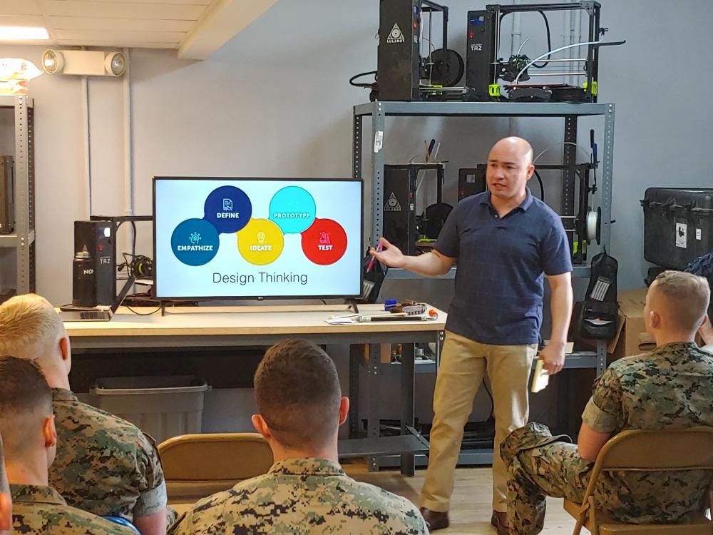 Um grupo de soldados assistindo uma apresentação sobre design thinking em uma televisão com uma pessoa de pé apresentando.