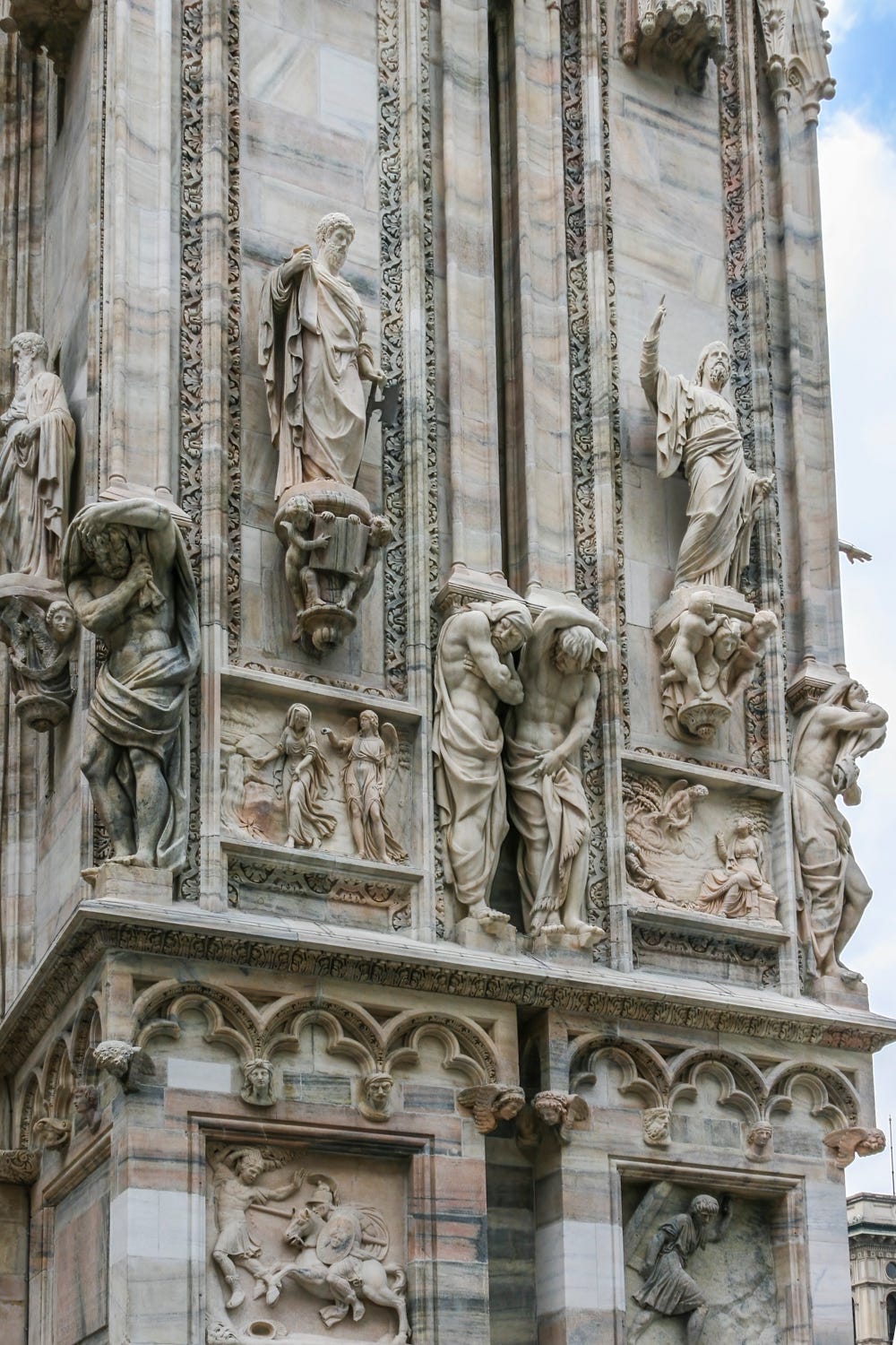 Detail of Milan Cathedral front facade. More than a dozen life-size statues plus a few smaller statues in bas-relief form. Blue, pink and white tones in the marble.